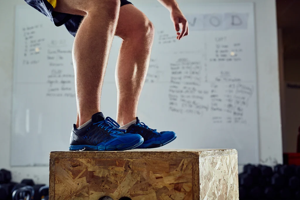 Primo piano delle gambe di un atleta che atterra su un box di legno durante un esercizio di salto in palestra