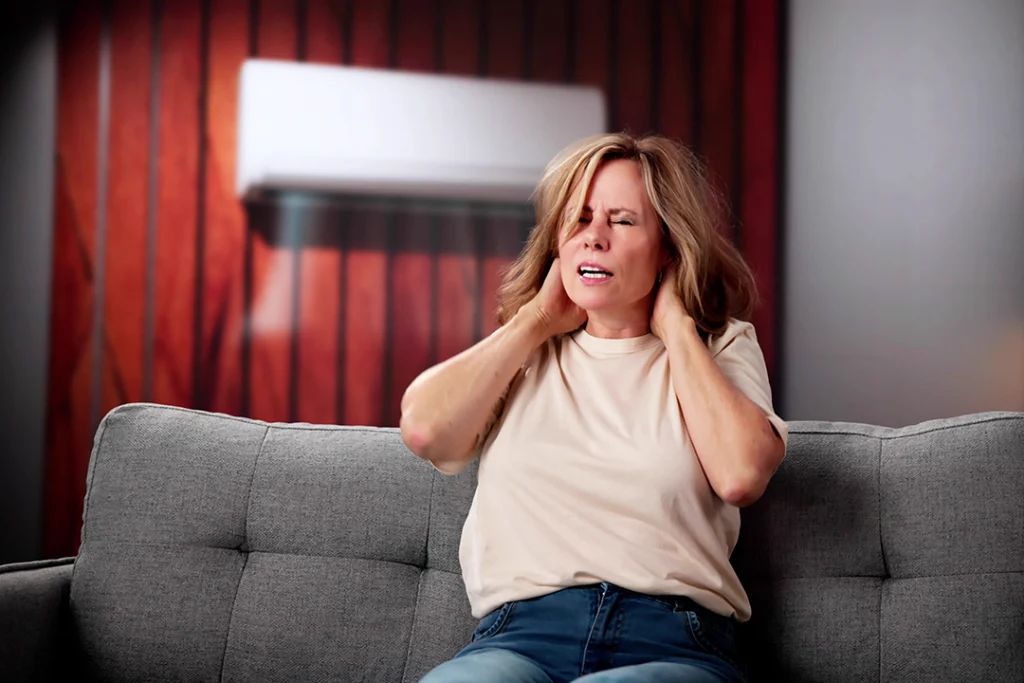 Donna seduta sul divano con espressione sofferente mentre si tiene il collo, sullo sfondo un condizionatore acceso. Sintomo di cervicalgia o dolore muscolare