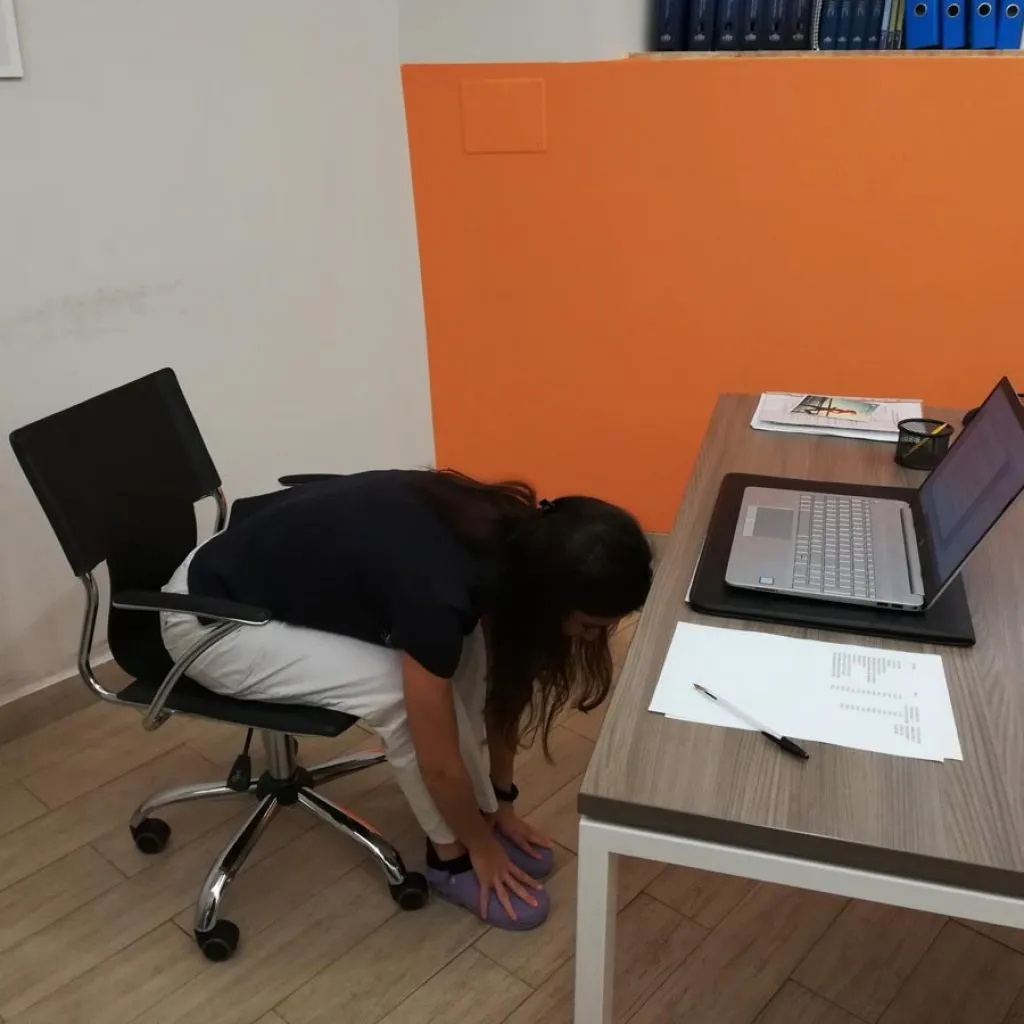 onna esegue esercizio di stretching alla scrivania piegandosi in avanti da seduta per toccare i piedi, attività utile per prevenire il mal di schiena in ufficio