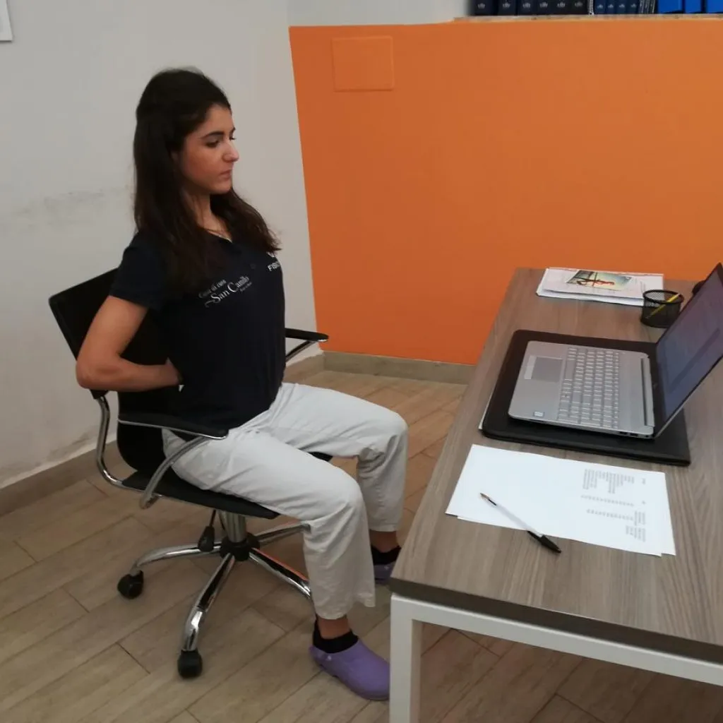 Donna seduta alla scrivania esegue esercizio per l’estensione lombare con le mani intrecciate dietro la schiena e le spalle aperte