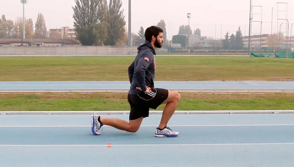 Persona che esegue un affondo su una pista all'aperto, indossando abbigliamento sportivo; sullo sfondo prato, alberi e edifici
