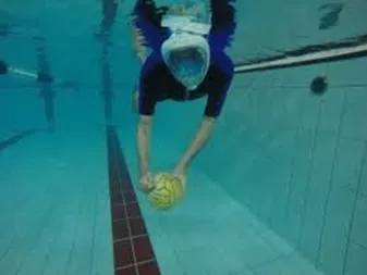 Persona immersa sott'acqua in piscina, intenta a raccogliere un pallone giallo dal fondo, con maschera e boccaglio, in posizione orizzontale