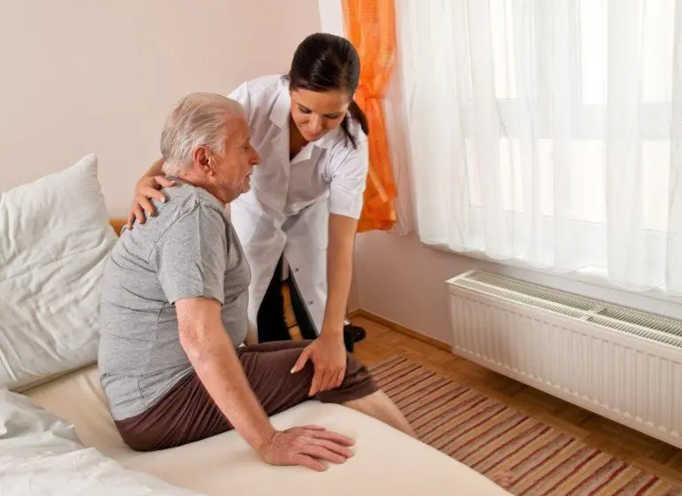 Anziano seduto nel letto e donna dal camice bianco che lo sorregge